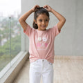 Niña con polo rosa de manga corta y diseño frontal Good Day con flores, combinado con pantalón blanco.